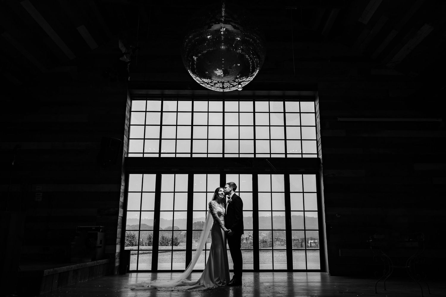 A dark industrial interior with large windows and beams silhouetted against bright natural light.