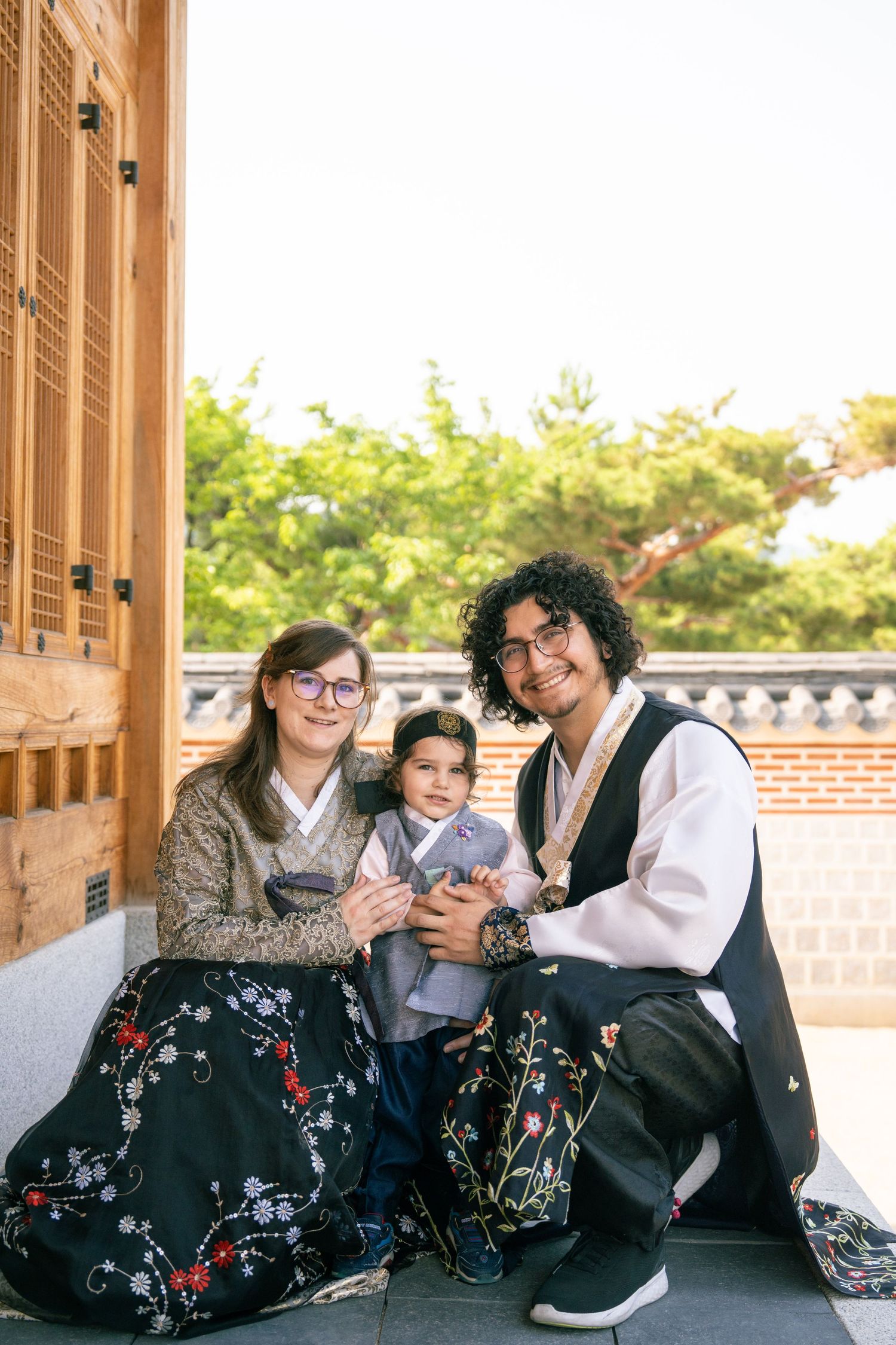 Three friends in traditional Korean hanbok dresses sit together on a wooden porch sharing a casual moment.
