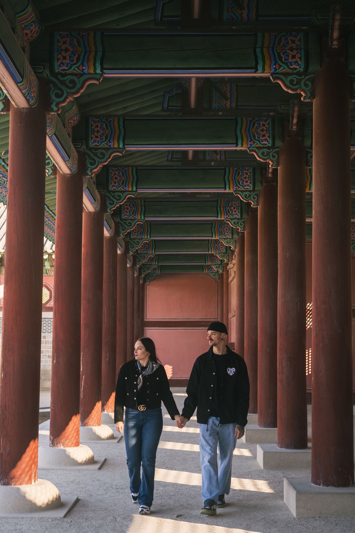 Couple walking hand in hand through ornate covered walkway with traditional Korean architecture and red pillars.