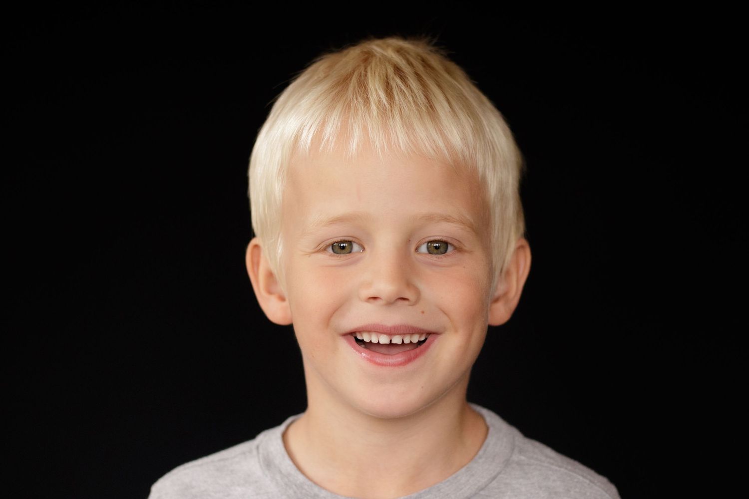 Happy blonde boy with a big smile wearing a gray shirt against a black background.