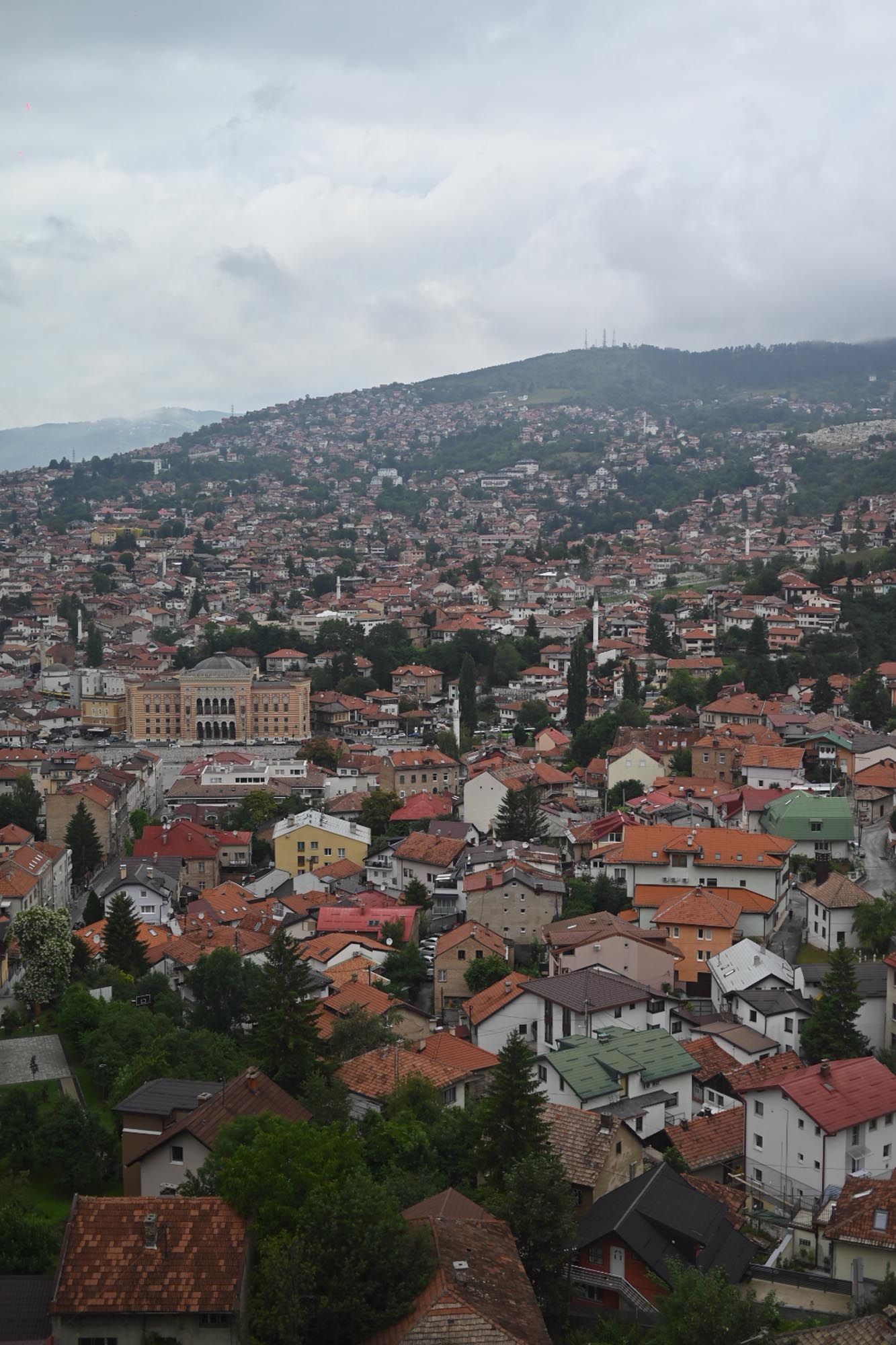 Panoramic cityscape view of Sarajevo with red-roofed buildings nestled in the valley surrounded by green hills.
