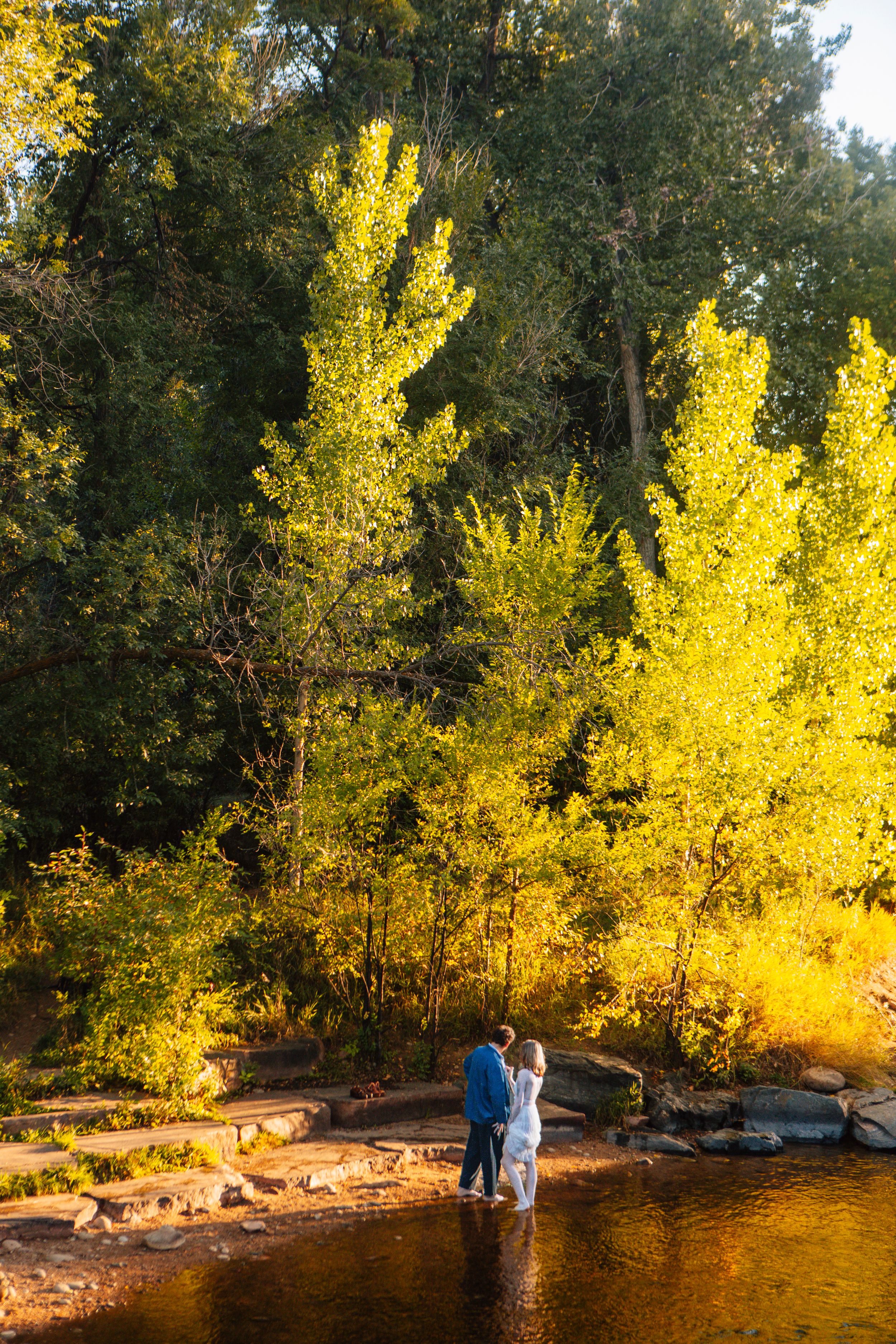 A couple stands at the edge of a lake surrounded by vibrant yellow autumn trees in golden afternoon light.