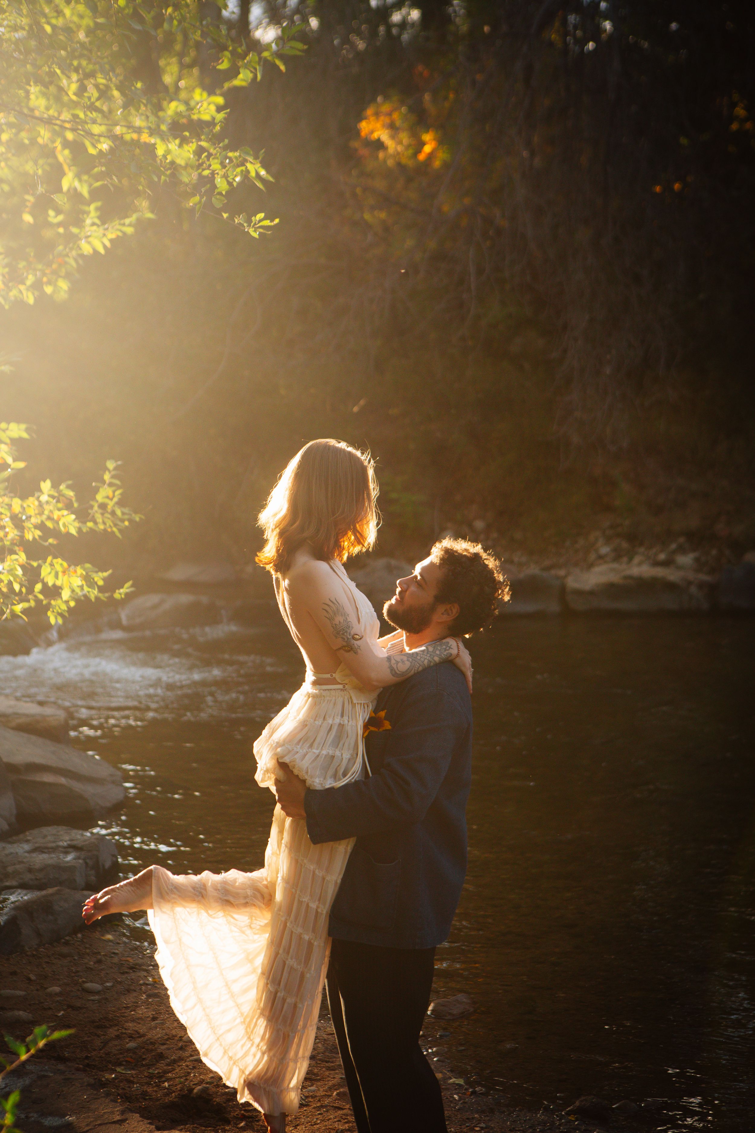 A romantic couple in formal attire dances together by a river at sunset with golden light filtering through trees.
