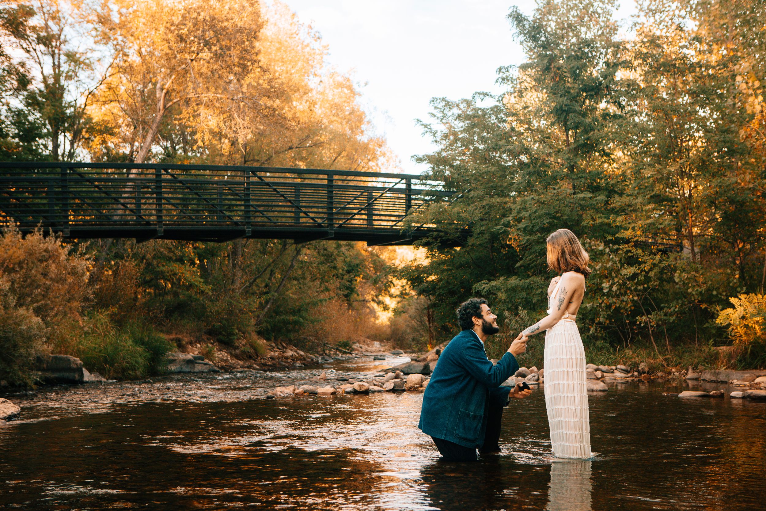 A couple stands on a rustic bridge during golden hour while capturing romantic autumn engagement photos by a river.
