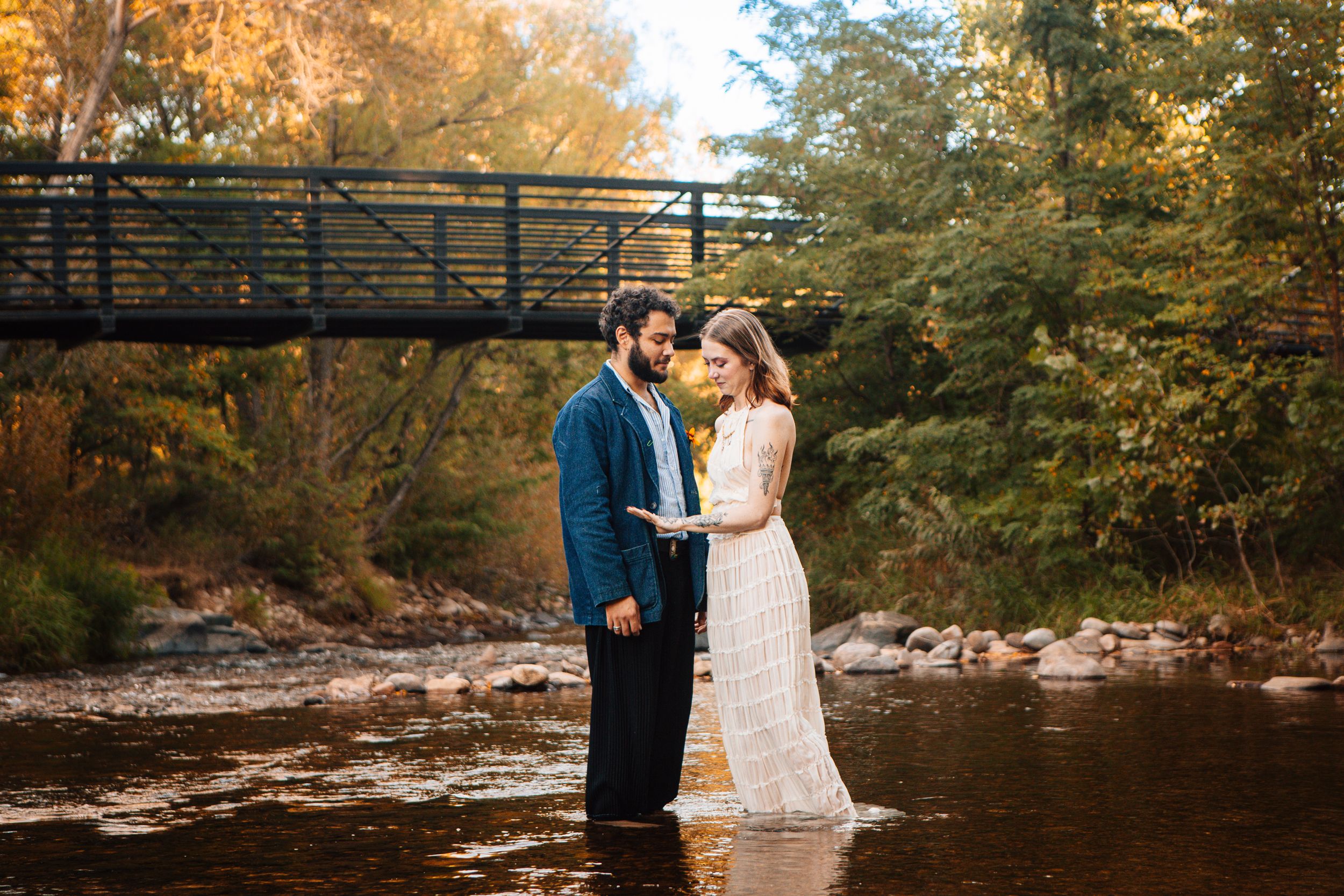 A couple shares an intimate moment on a rustic wooden bridge surrounded by autumn foliage and flowing water.