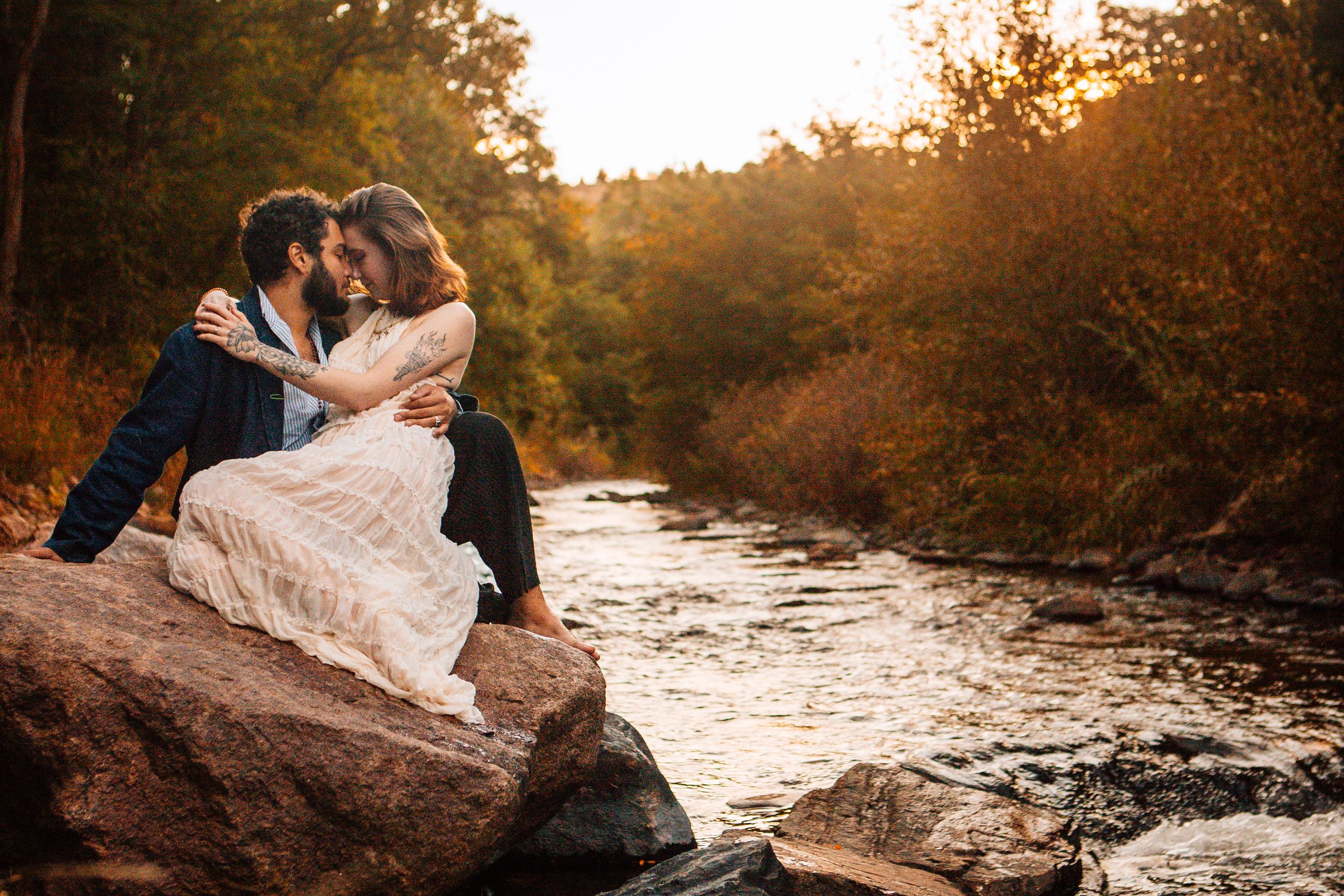 A couple in wedding attire sits on a large rock beside a flowing river at sunset in an autumn forest setting.