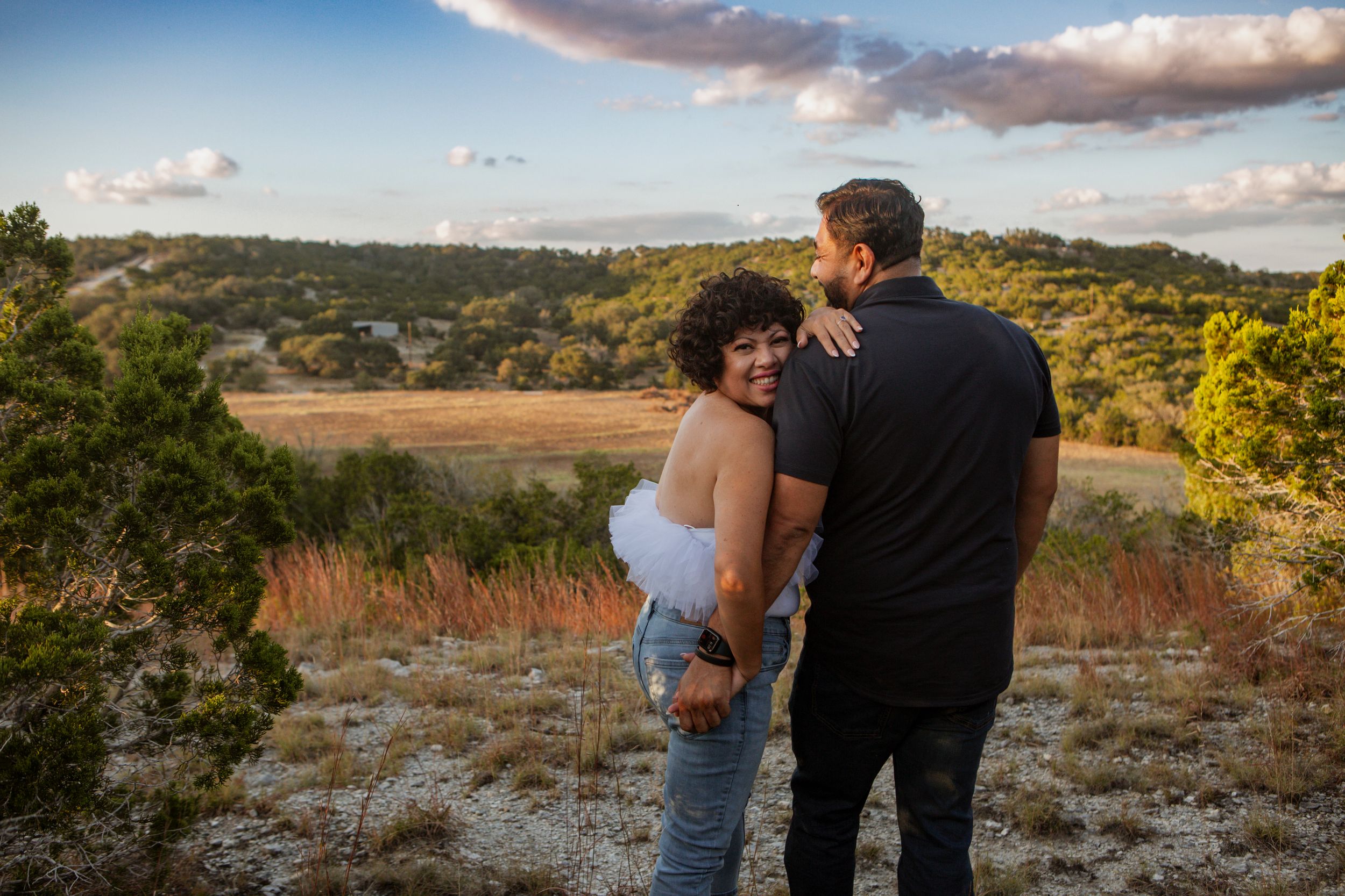 Couple stands together on hilltop during golden hour with scenic mountain landscape in background.