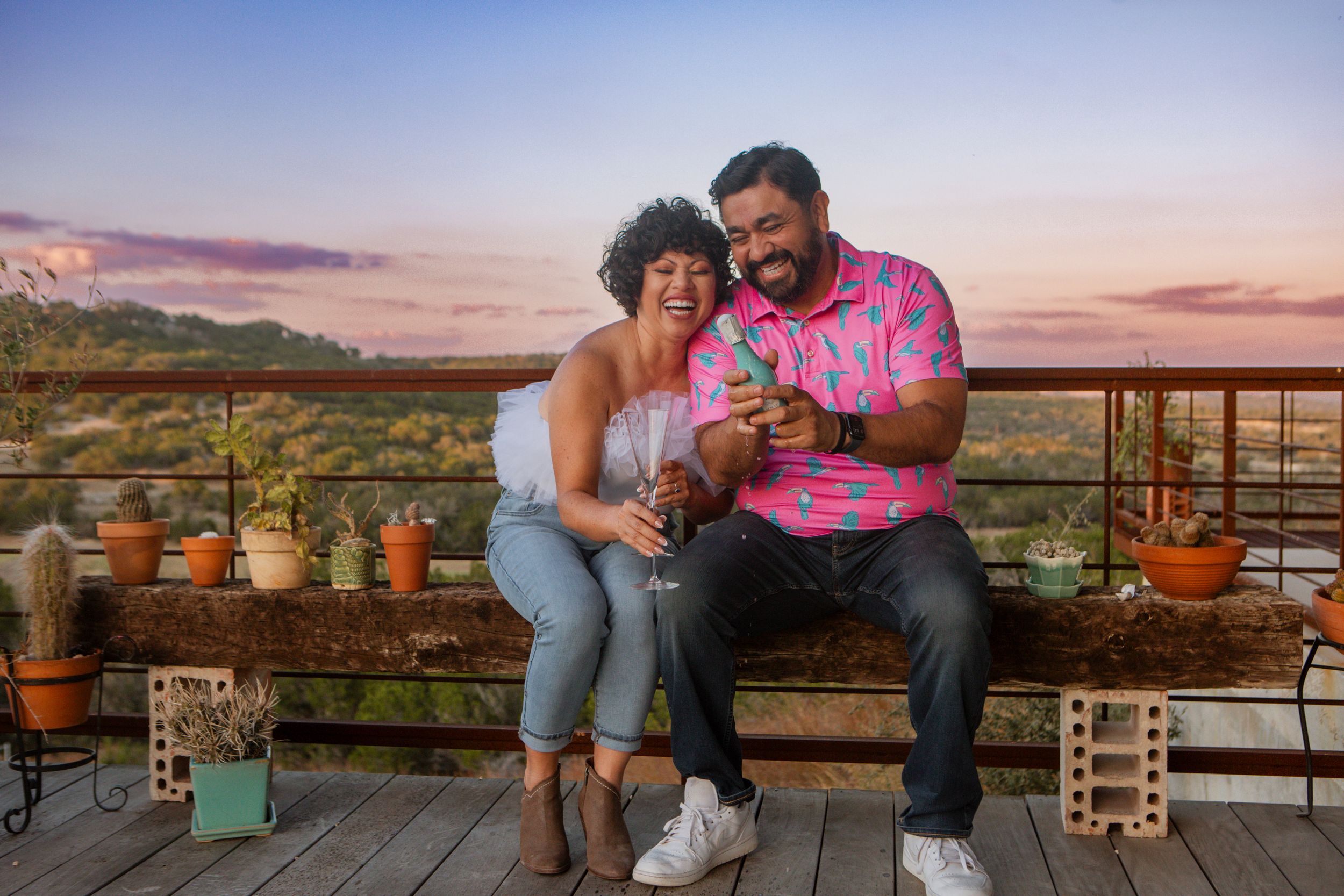 Couple sitting on outdoor patio at sunset sharing drinks and looking at phone with desert mountains in background.