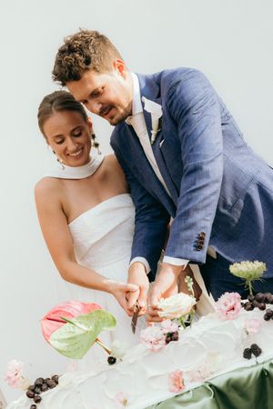 Couple cutting their wedding cake together decorated with pink flowers and anthurium leaves.