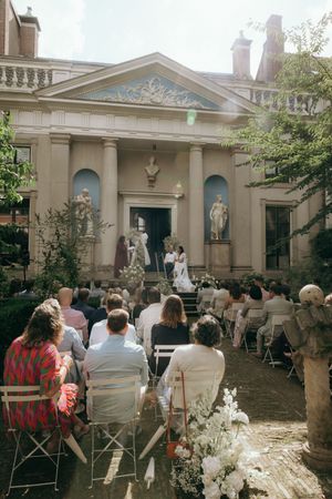 Outdoor wedding ceremony with guests seated in folding chairs facing a historic building with columns.
