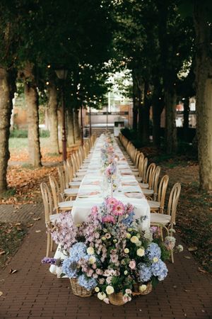 Long outdoor dining table setup with floral arrangements under a tree-lined path.