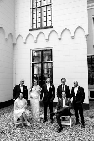 Wedding party poses in formal attire against a white building with gothic windows.