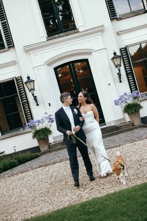 Wedding couple walks along a gravel path outside a white building with their dog.