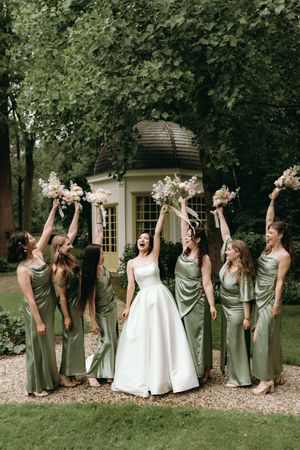 Wedding party in sage green dresses and white gowns posing playfully outdoors.