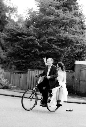 Black and white image of a couple joyfully riding a bicycle together on a residential street.