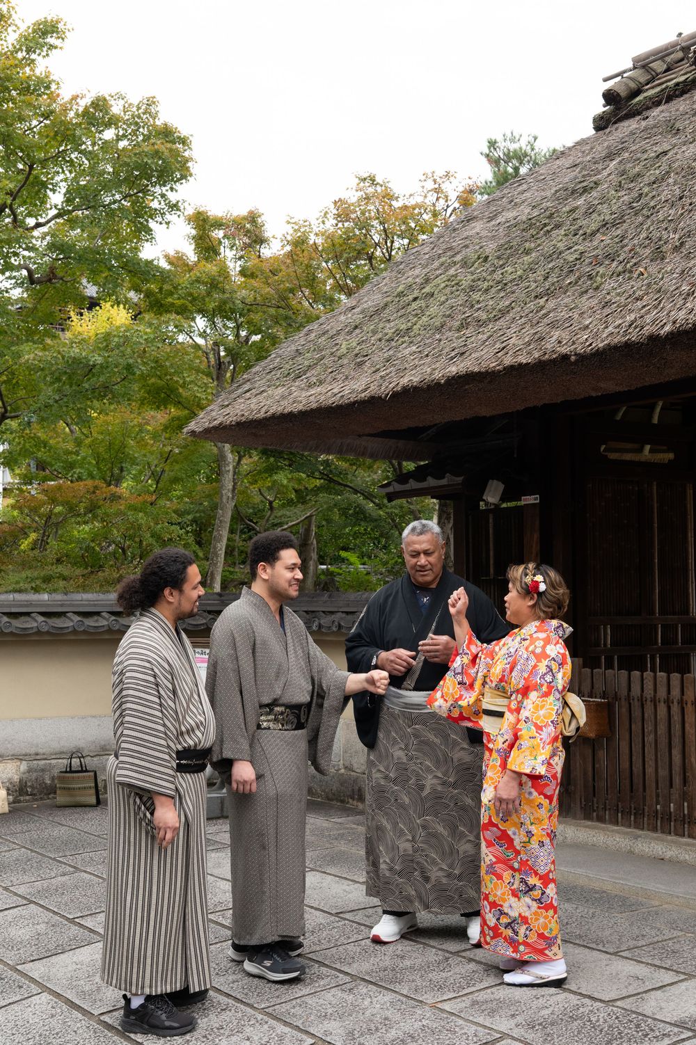 Family of four dressed in traditional Japanese kimono converse outside thatched-roof Kyoto building.