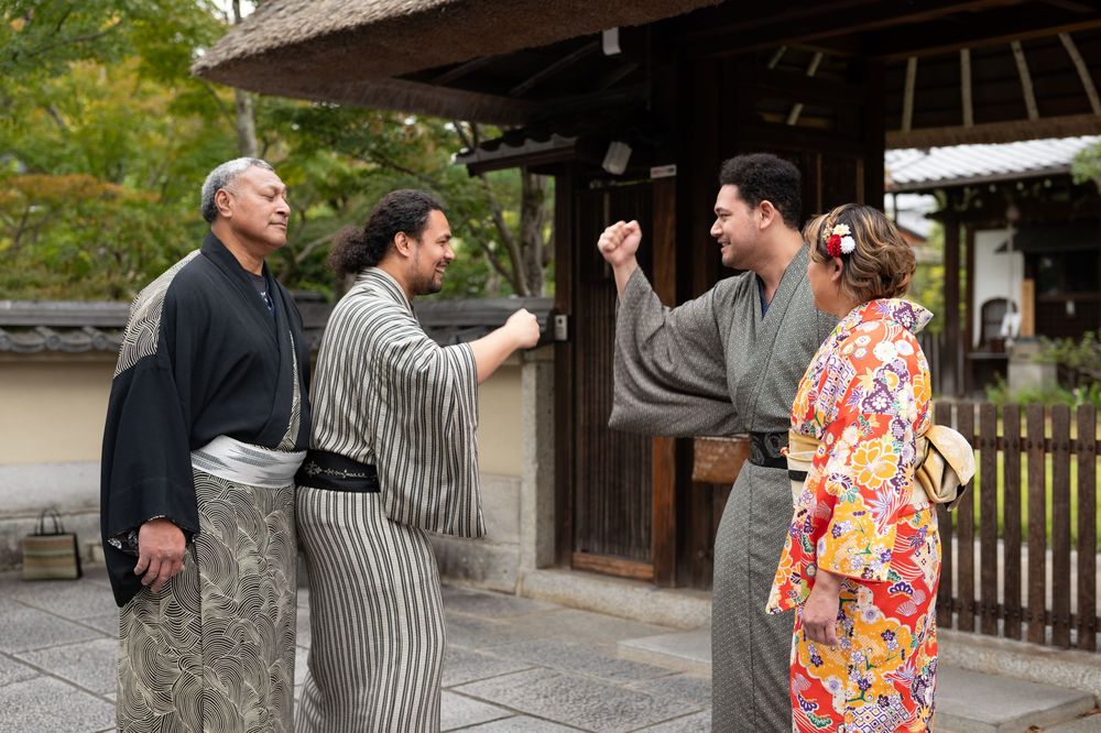 Four people wearing kimono laughing and greeting each other outside a Japanese temple gate.