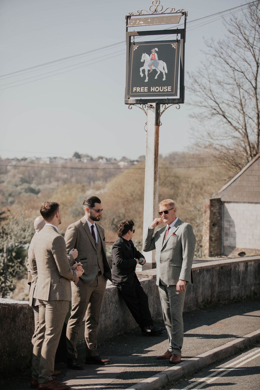 Groomsmen in tan suits socializing outside a Free House pub sign on a sunny wedding day.