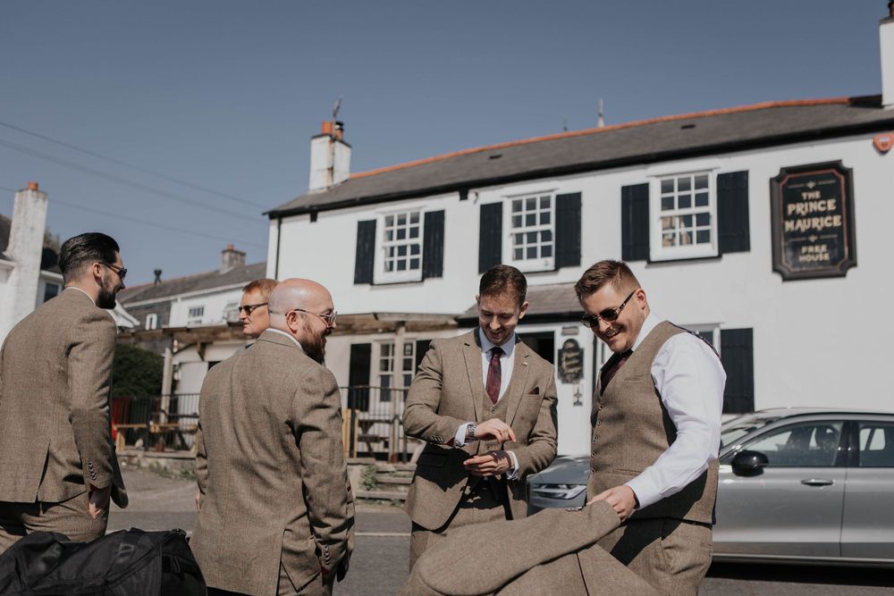 Groomsmen in tweed suits gathered outside a white pub building examining wedding details.