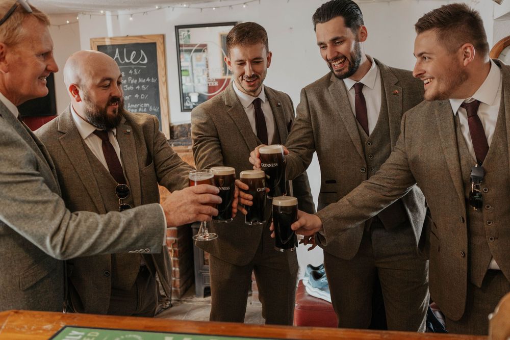 Five groomsmen in brown suits toasting with pints of Guinness at a pub bar.