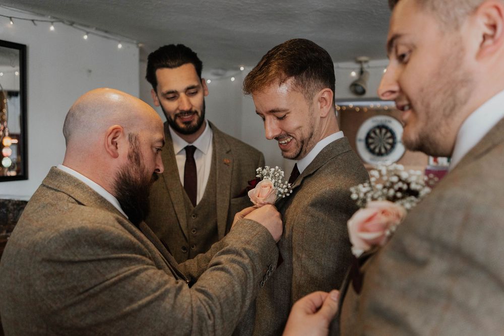 Groomsmen in brown tweed suits pinning a floral boutonniere on the groom inside a pub.
