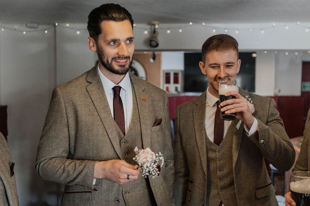 Two groomsmen in brown tweed suits smiling, one drinking a pint of Guinness at a pub.