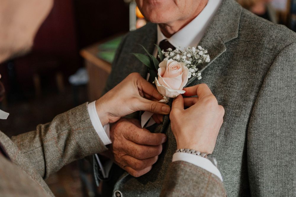 Close-up of hands pinning a blush pink rose boutonniere onto a grey tweed suit lapel.