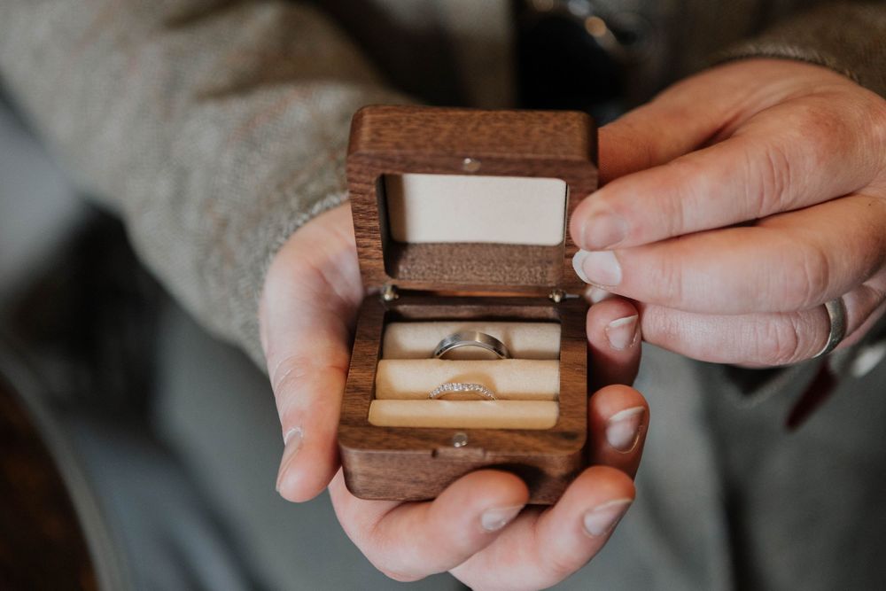 Hands holding an open wooden ring box containing two wedding bands on a cream velvet cushion.