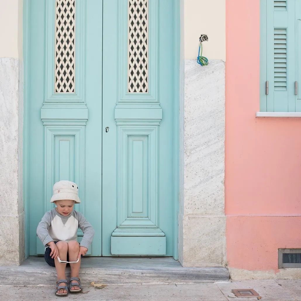 Turquoise painted wooden doors line a pink wall in a historic neighborhood with someone sitting on a doorstep.
