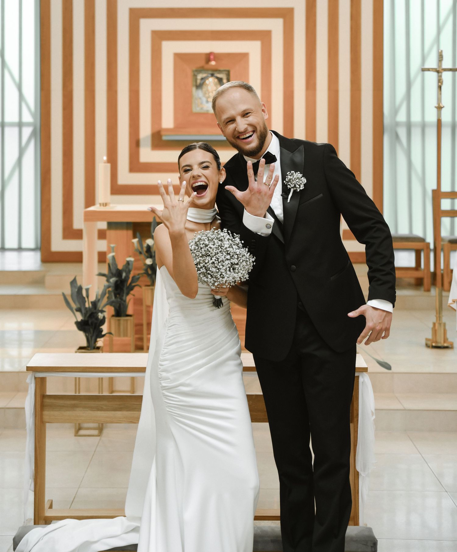Newlyweds and wedding party share joyful embraces in a church with geometric orange walls.