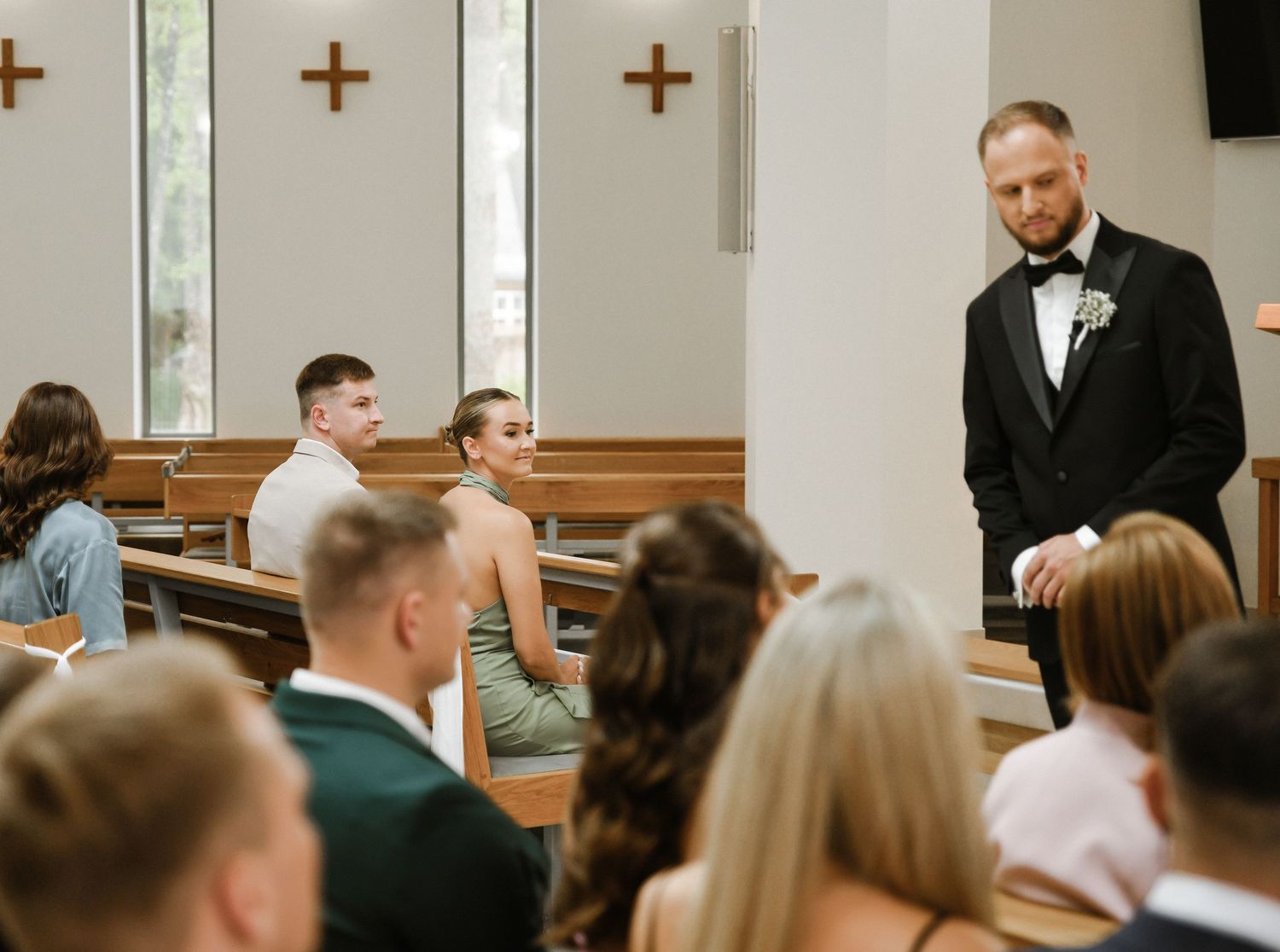 Wedding ceremony taking place in a bright church with wooden crosses on the wall and guests seated in pews.