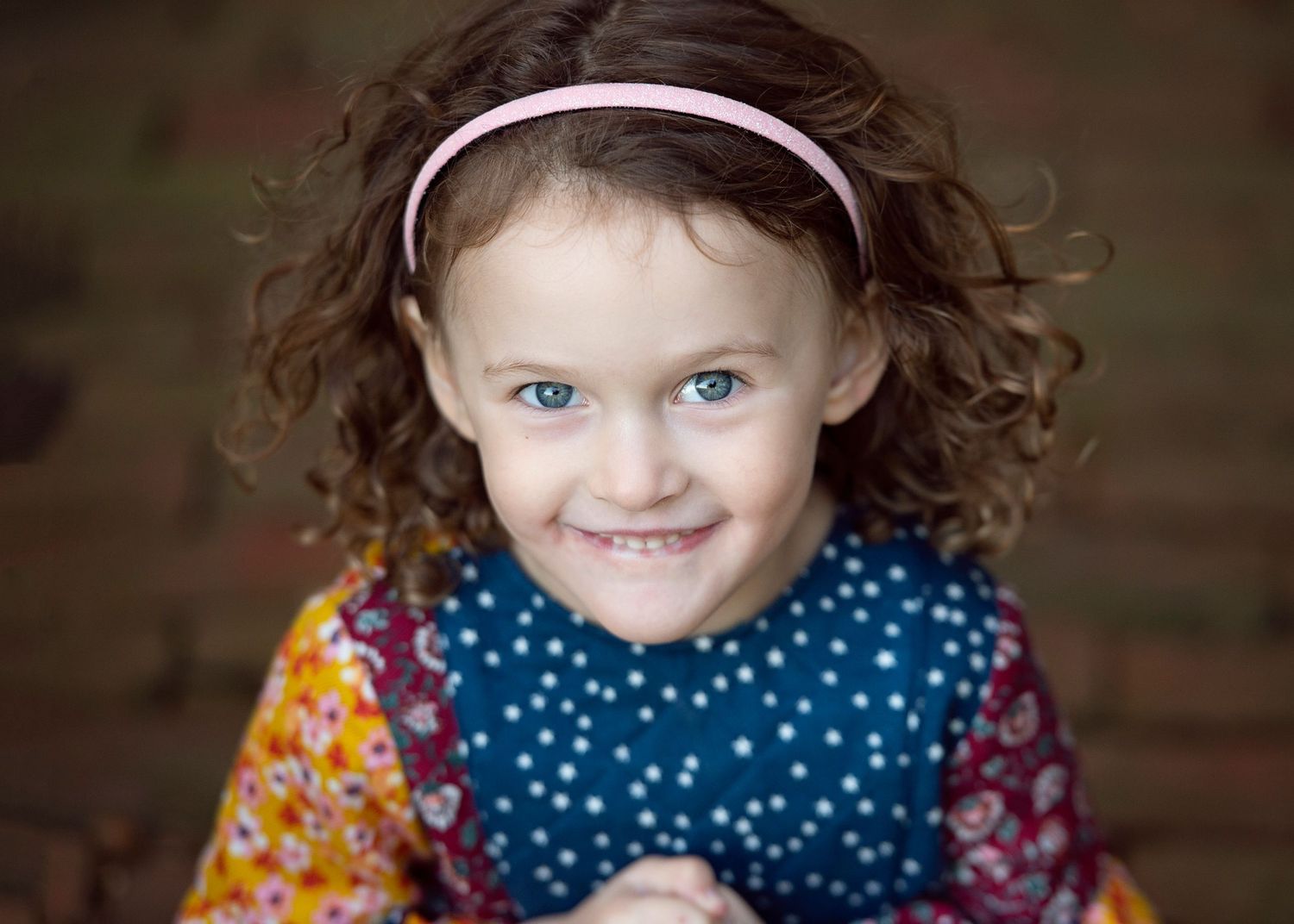 Smiling young girl with curly brown hair and blue eyes wearing a colorful floral outfit and pink headband.
