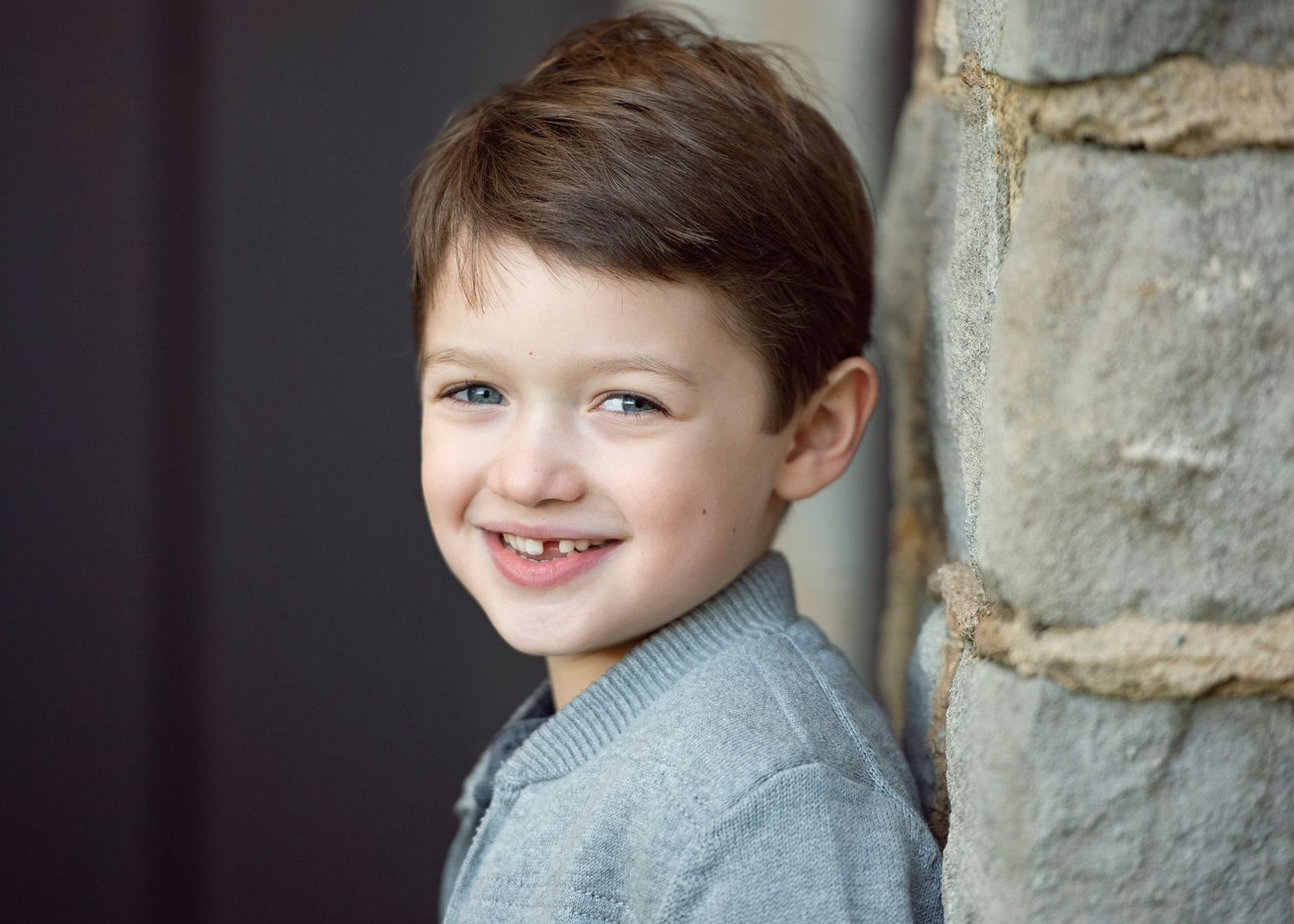 A cheerful young boy with brown hair smiles while leaning against a stone wall, wearing a gray sweater.