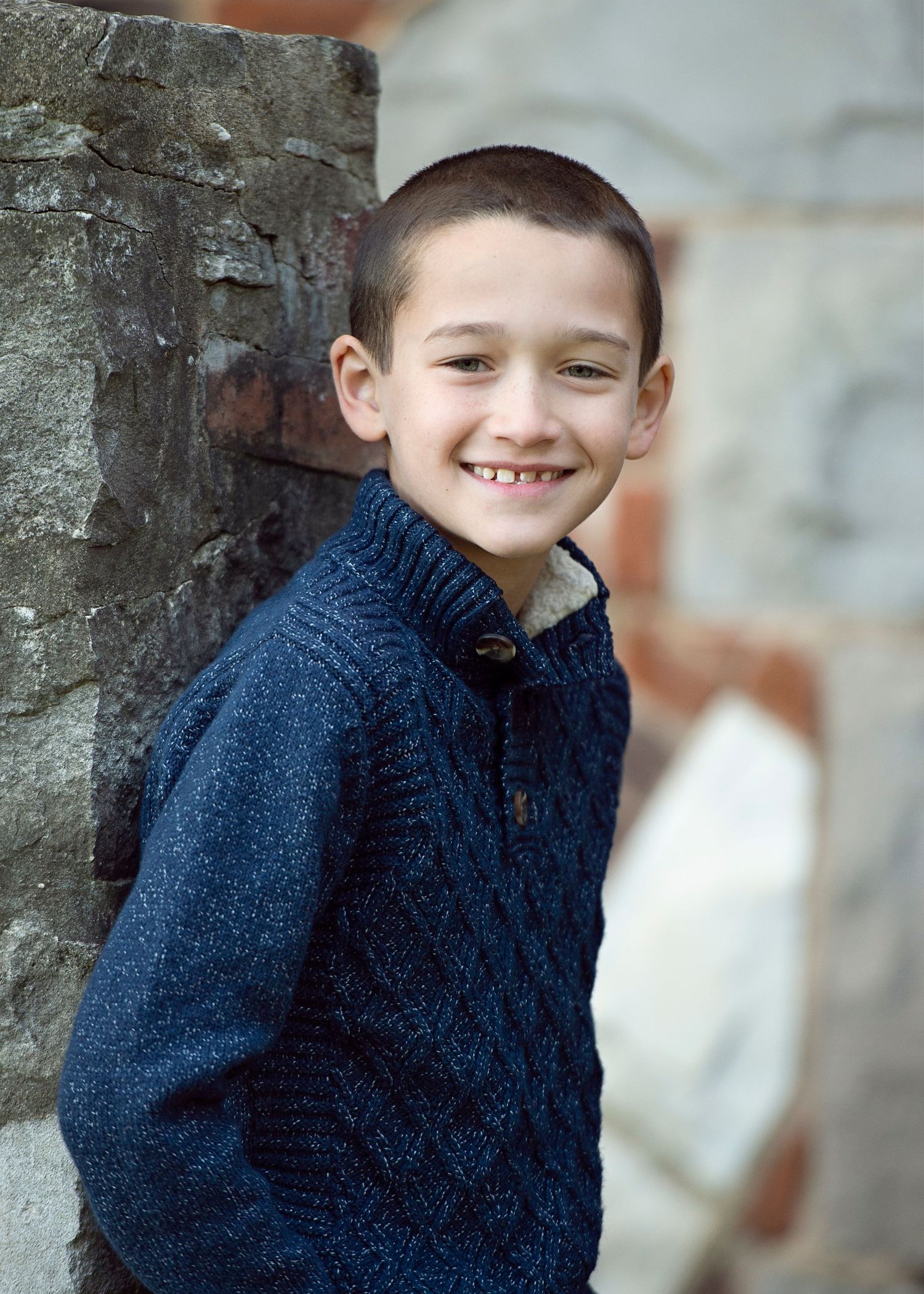 Smiling young boy in a navy blue cable-knit sweater leaning against a stone wall outdoors.