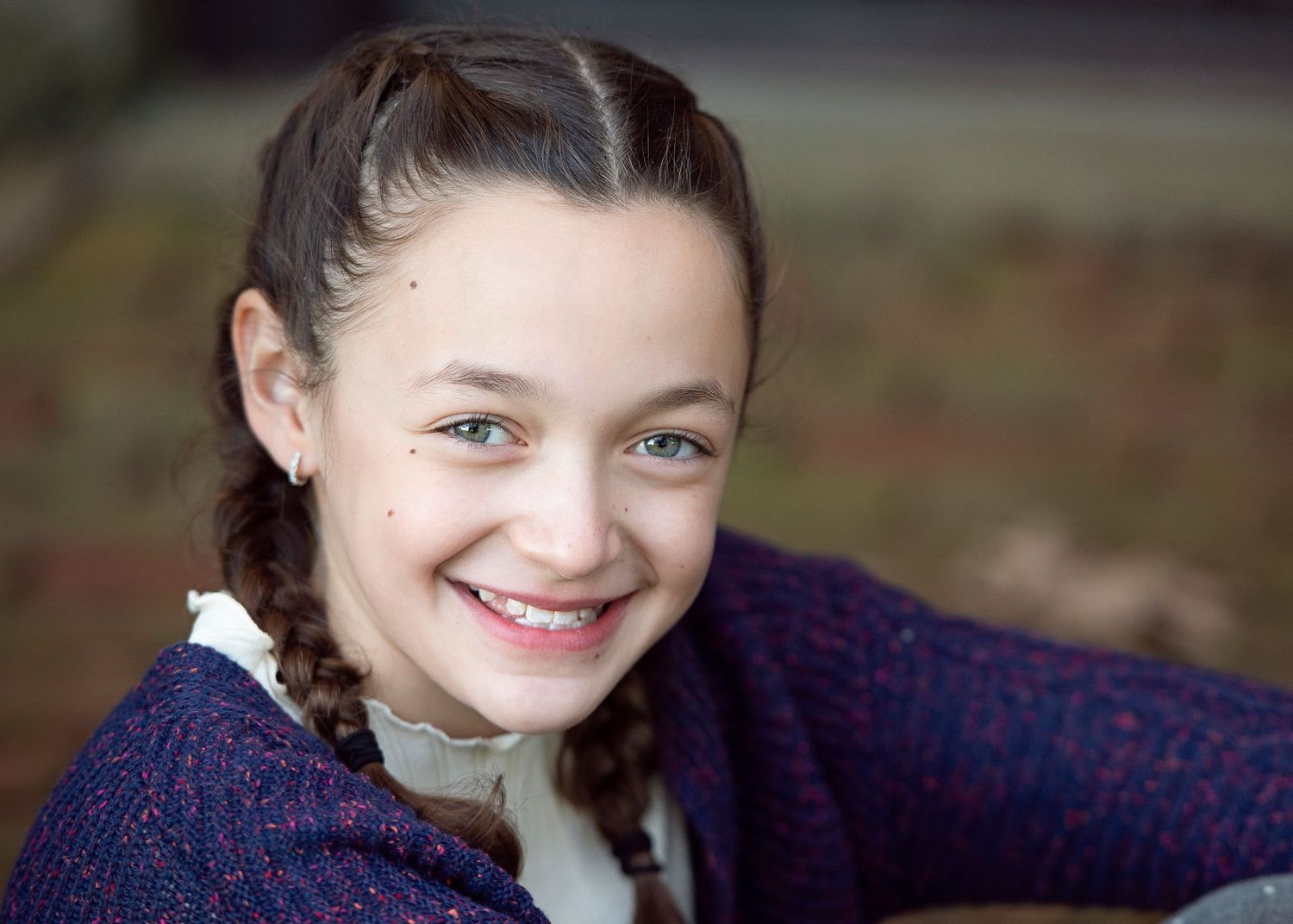 Smiling young girl with braided hair wearing a blue cardigan outdoors in a candid portrait photo.