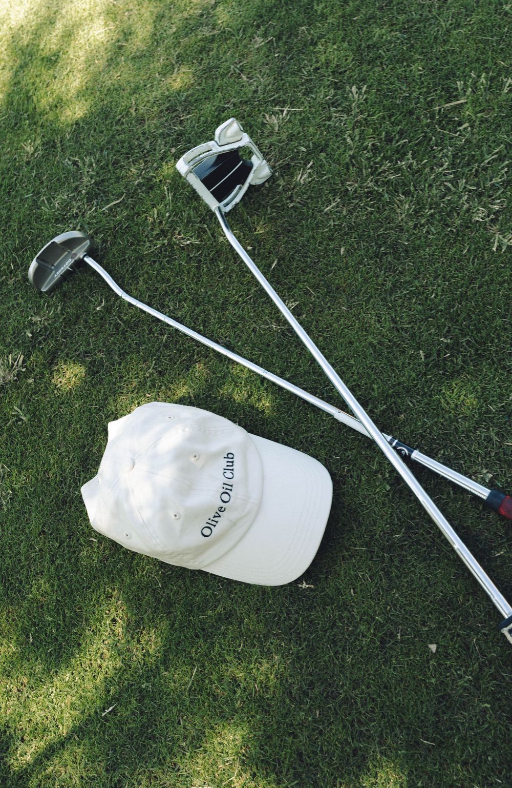Golf clubs and a white cap lying on green grass in dappled sunlight.