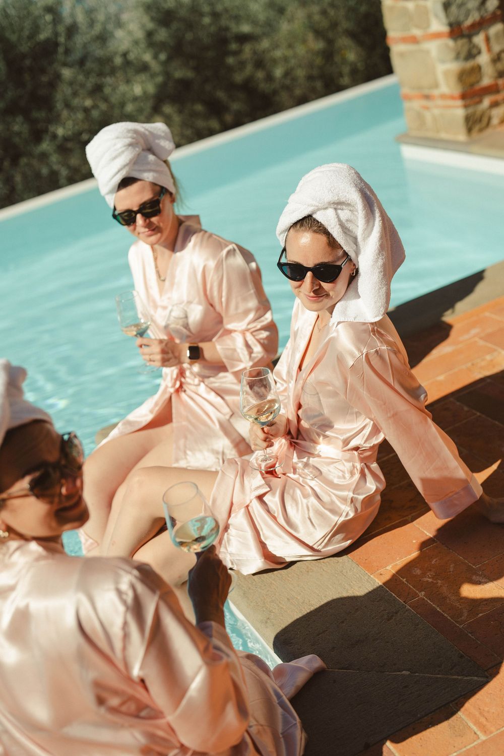Bridesmaids in pink silk robes and hair towels lounging by a pool.