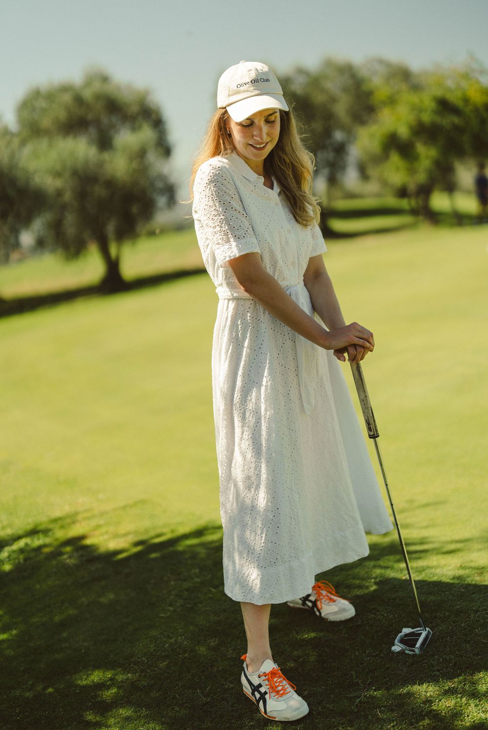 A person in a white dress and baseball cap poses with a golf club on a sunny course.