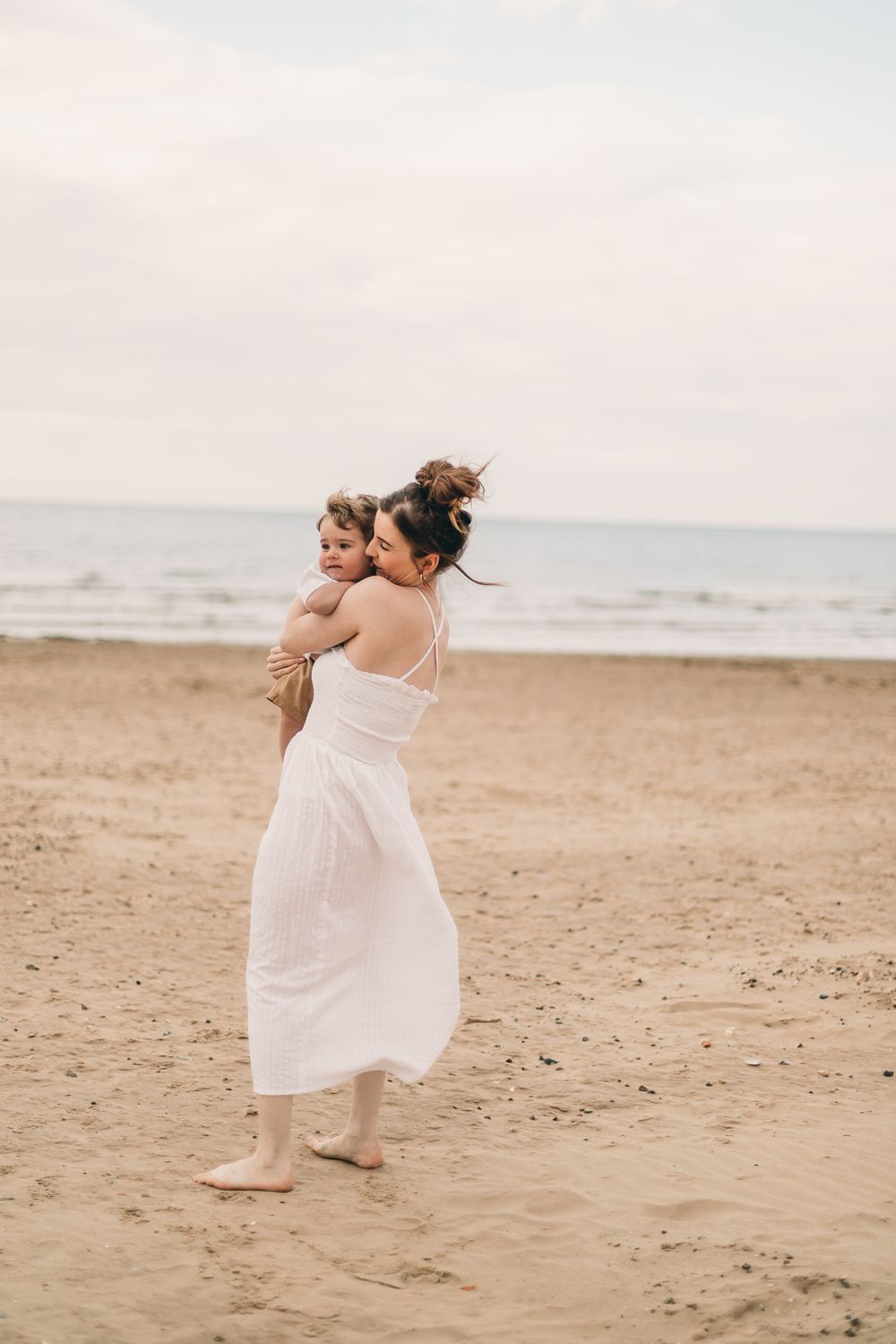 A mother in a white dress holds her young child on a sandy beach with calm ocean waves in the background.