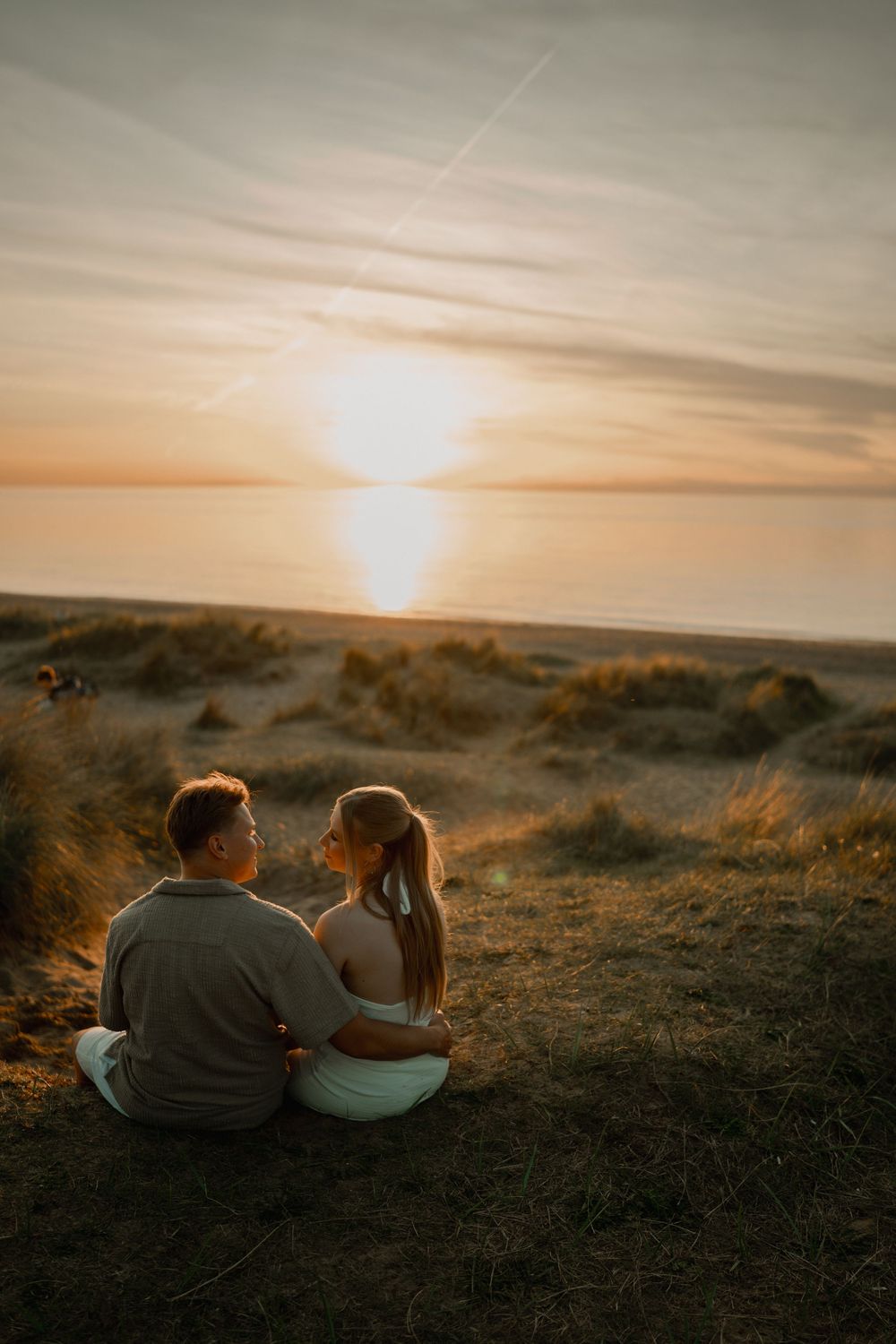 Couple sitting together on coastal dunes watching a golden sunset over the ocean.