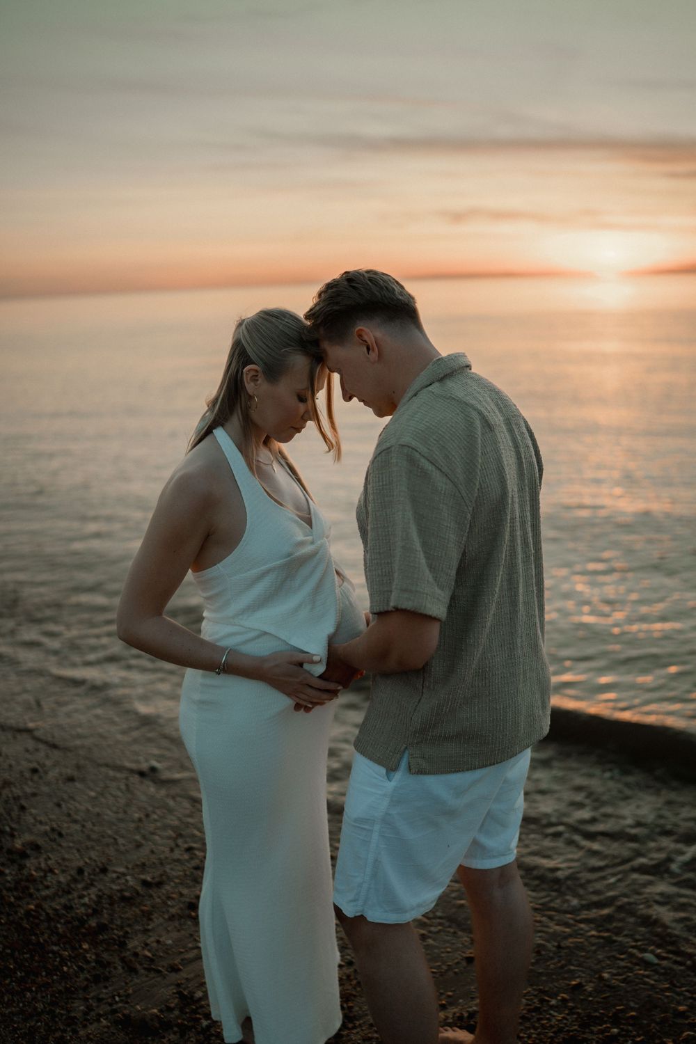 Couple sharing a tender moment at sunset by the ocean, woman in white dress cradling pregnancy bump.