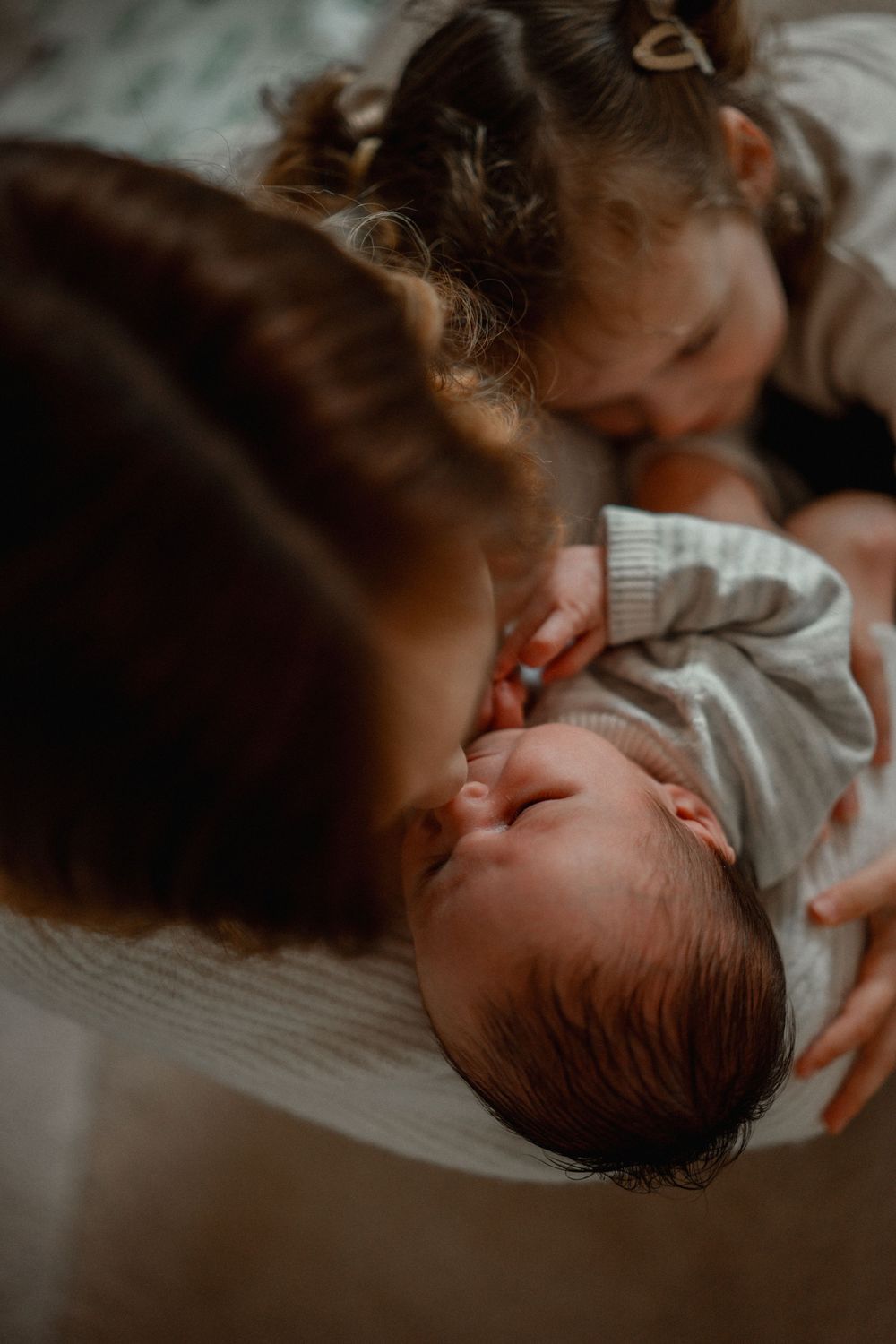 Mother breastfeeding newborn baby while older toddler sister leans in to give a gentle kiss.
