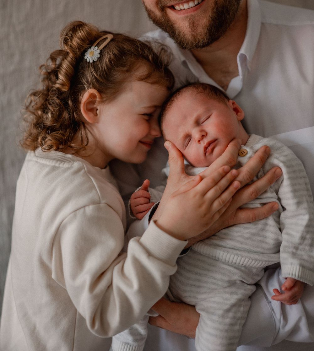 Father holds sleeping newborn while young curly-haired girl lovingly nuzzles baby sibling in tender family moment.