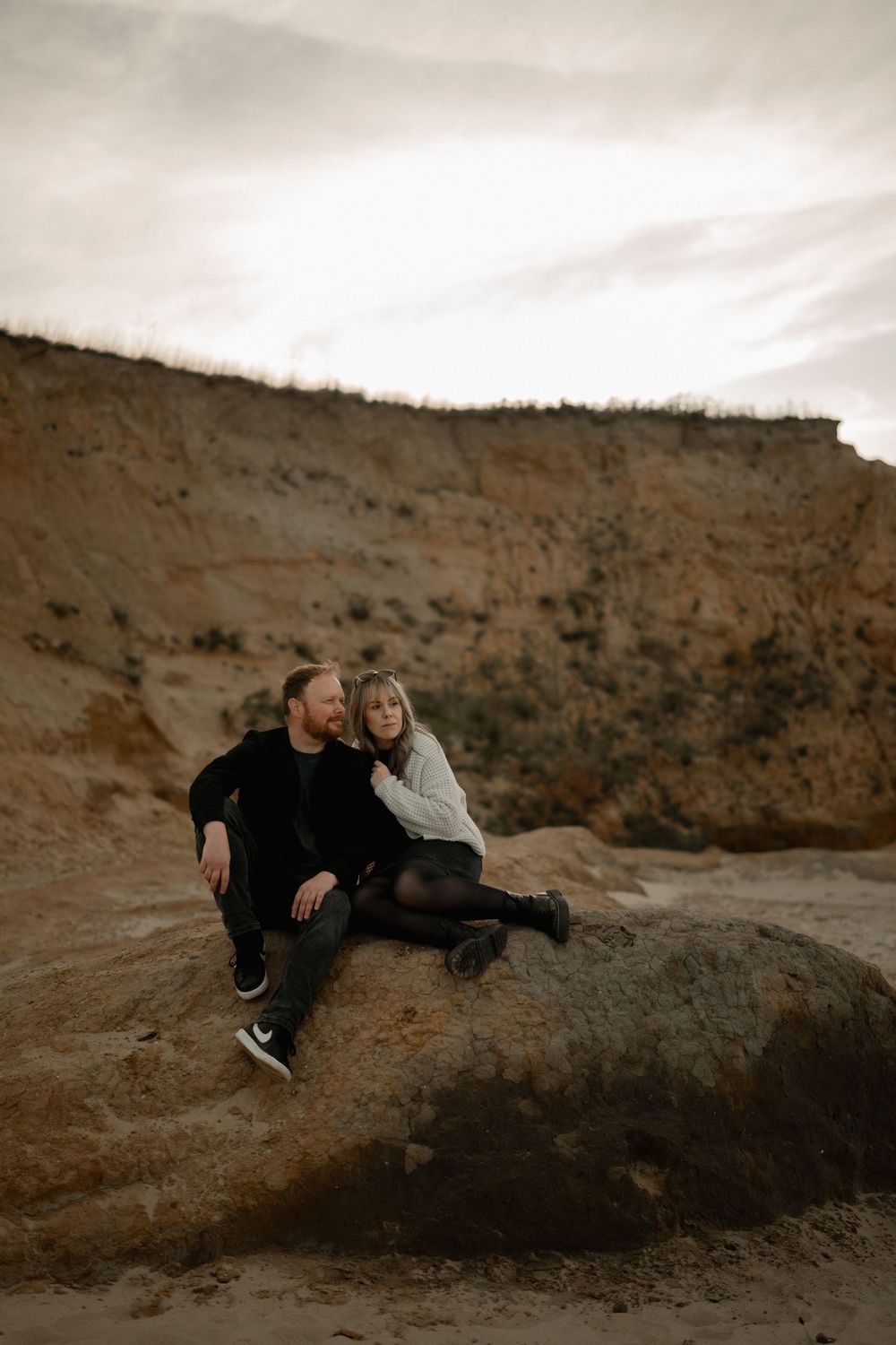 A couple sits closely on a rock in a sandy canyon, sharing a tender moment against a moody, overcast sky.