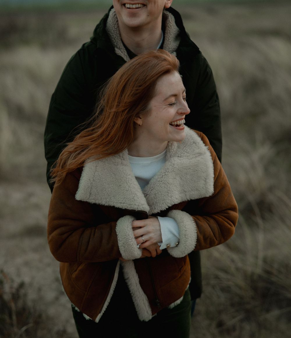 A smiling redhead woman in a brown shearling jacket embraces a man outdoors in a grassy field.
