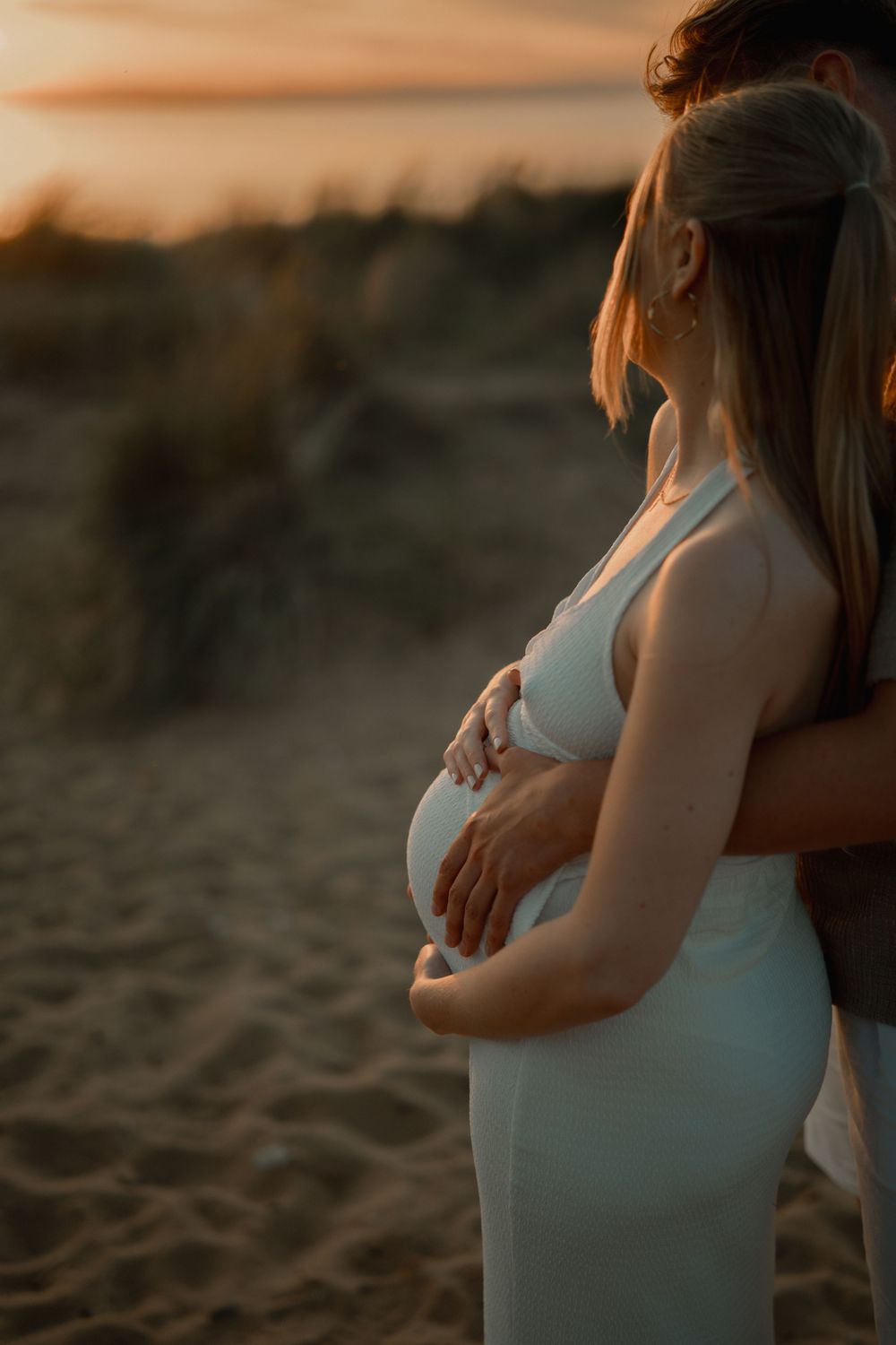 Pregnant woman in white dress cradled by partner at golden sunset on sandy beach.