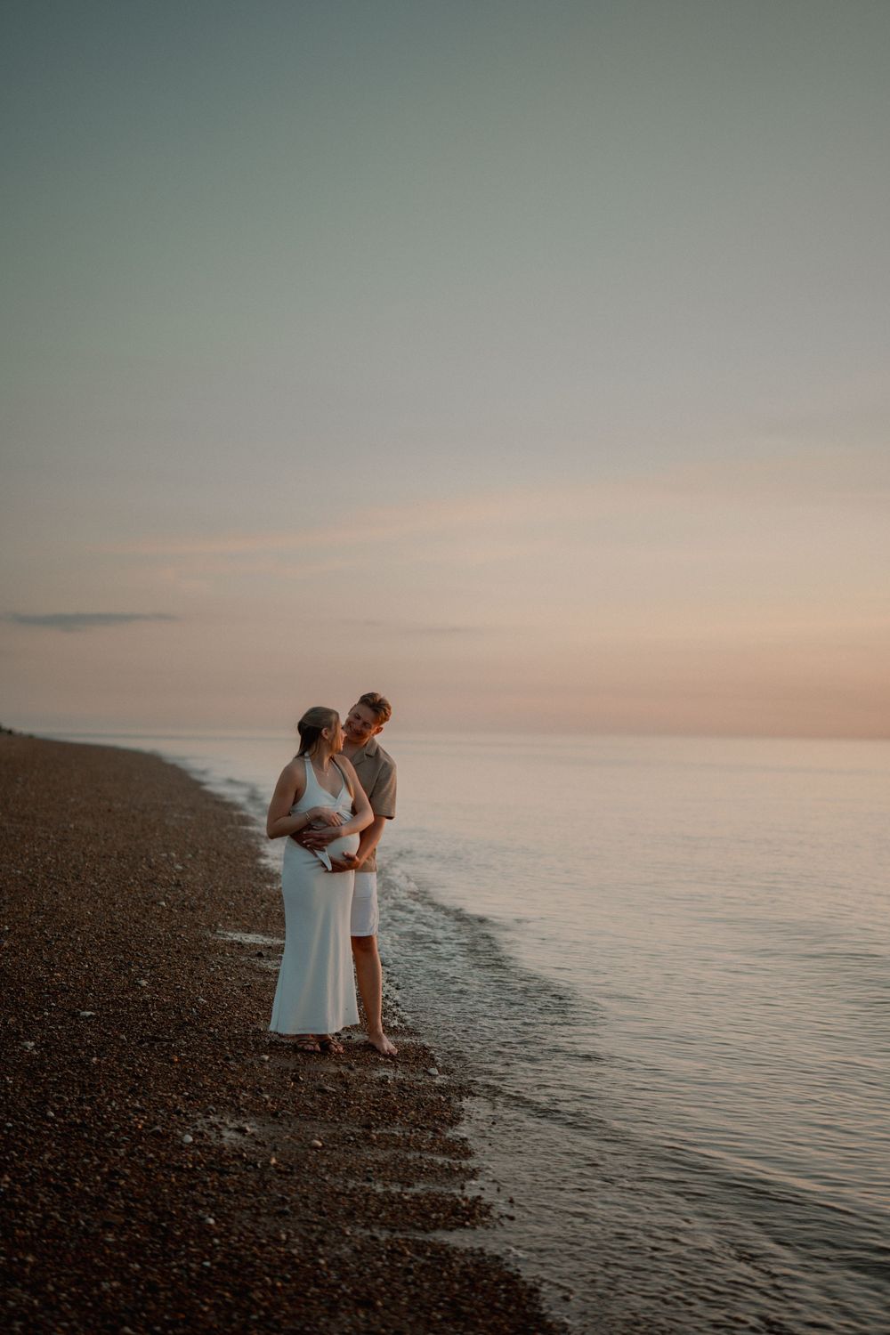 Couple standing on a pebble beach at sunset, woman in white dress with baby bump, embraced by partner.