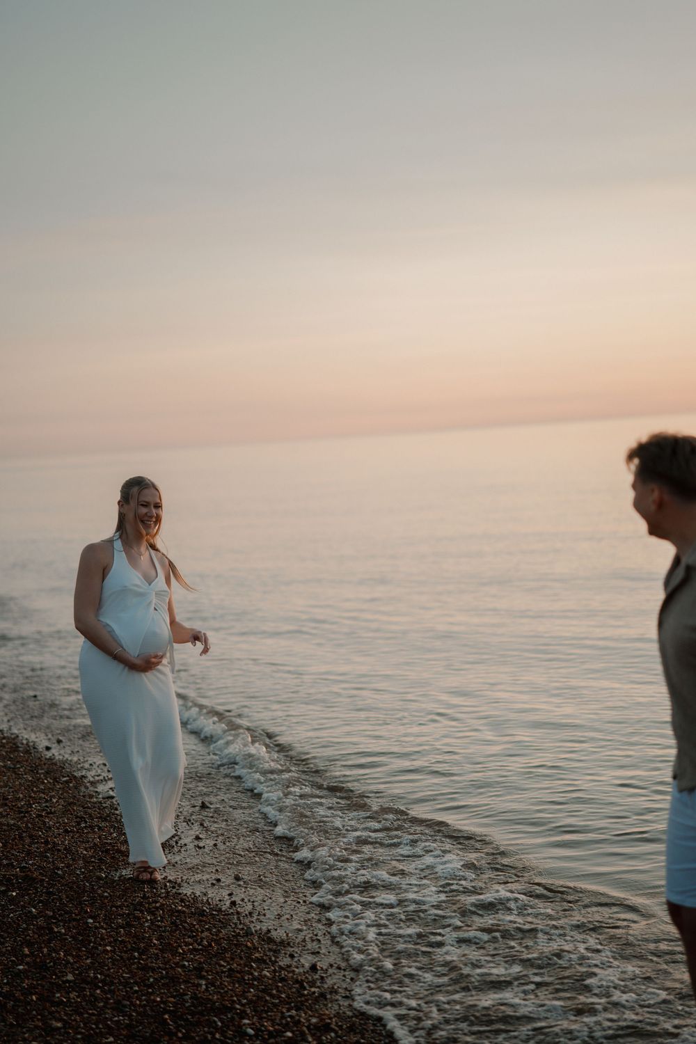 Pregnant woman in white dress smiling on a pebble beach at sunset with a man standing nearby.