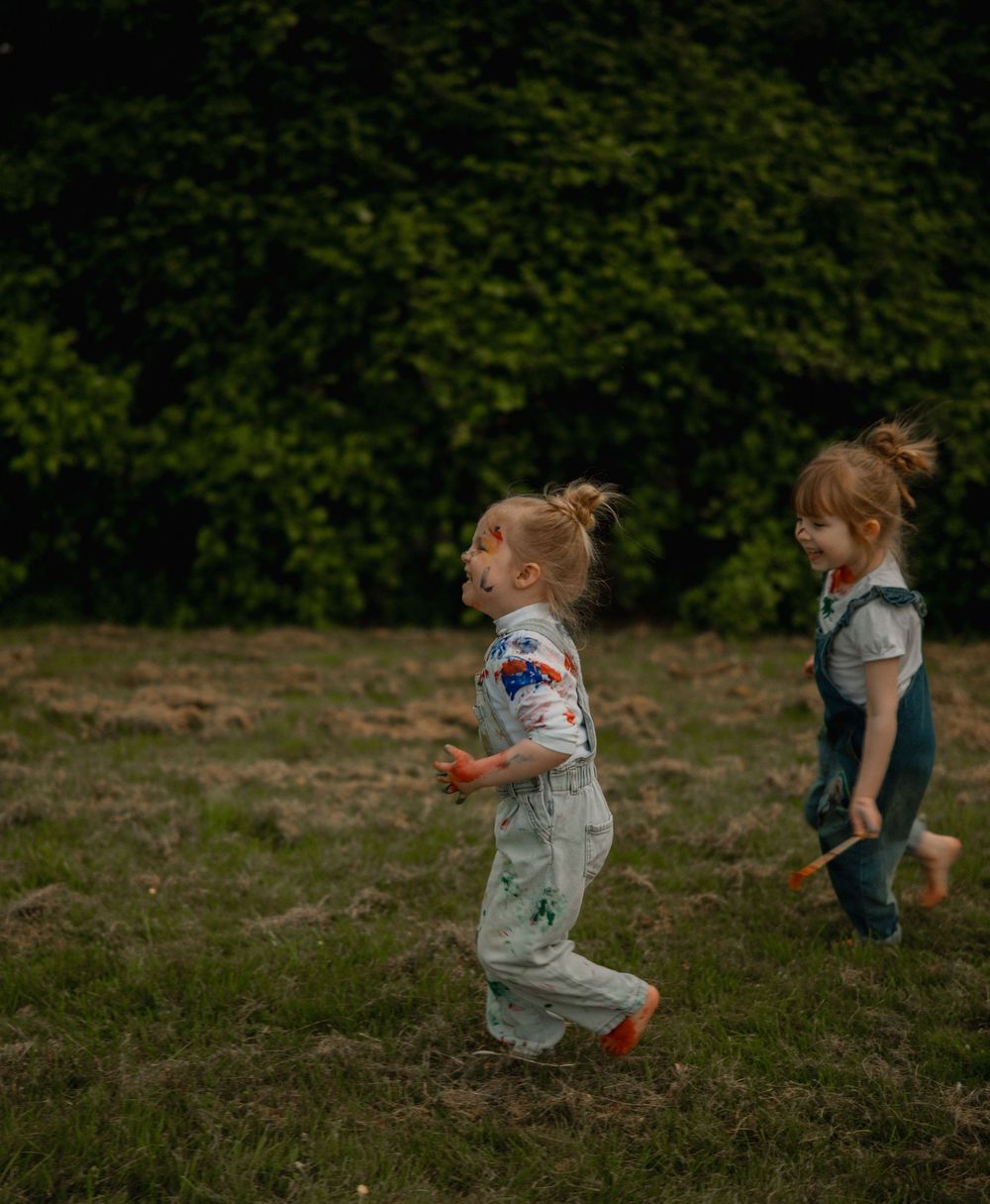 Two young girls running and playing outdoors in a grassy field surrounded by lush green trees.