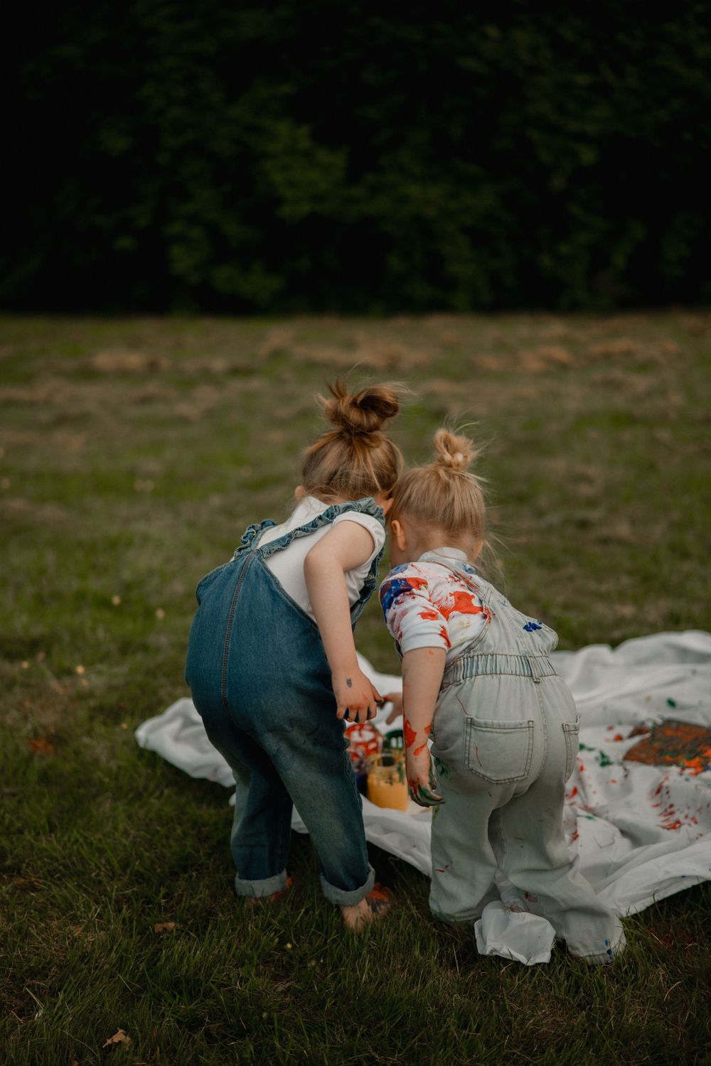 Two toddler girls in denim overalls play together on a white blanket in a lush green park.